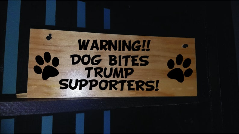 Puede incluir: Un letrero de madera con texto negro que dice "WARNING!! DOG BITES TRUMP SUPPORTERS!" con una huella de pata negra a cada lado.