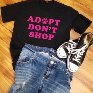 K&ouml;nnte beinhalten: Ein schwarzes T-Shirt mit dem pinkfarbenen Text "ADOPT DON'T SHOP", kombiniert mit blauen Jeans und schwarzen Canvas-Sneakern mit wei&szlig;en Schn&uuml;rsenkeln. Die Artikel sind auf einer Holzoberfl&auml;che angeordnet.