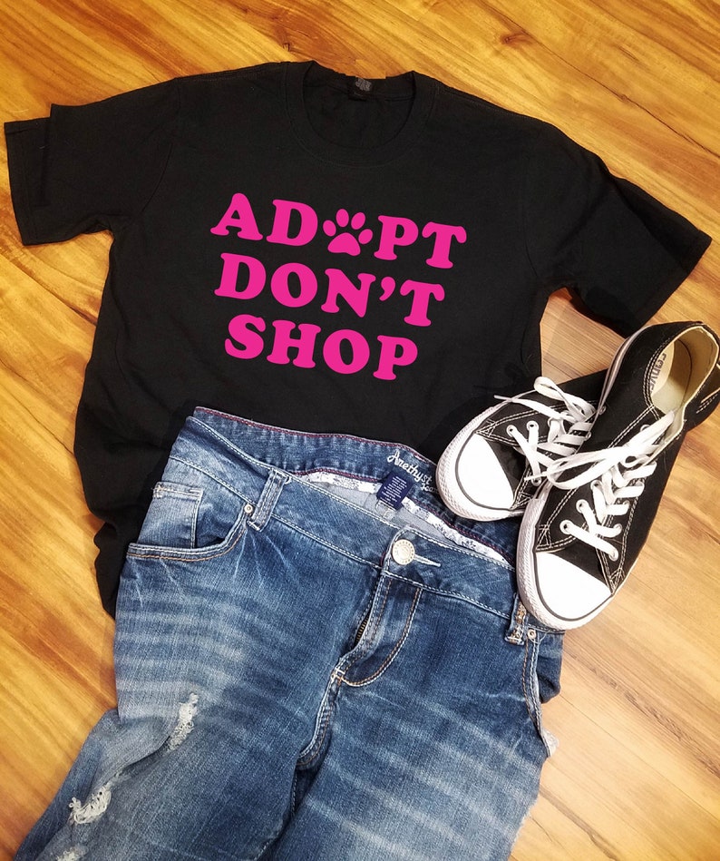 K&ouml;nnte beinhalten: Ein schwarzes T-Shirt mit dem pinkfarbenen Text "ADOPT DON'T SHOP", kombiniert mit blauen Jeans und schwarzen Canvas-Sneakern mit wei&szlig;en Schn&uuml;rsenkeln. Die Artikel sind auf einer Holzoberfl&auml;che angeordnet.