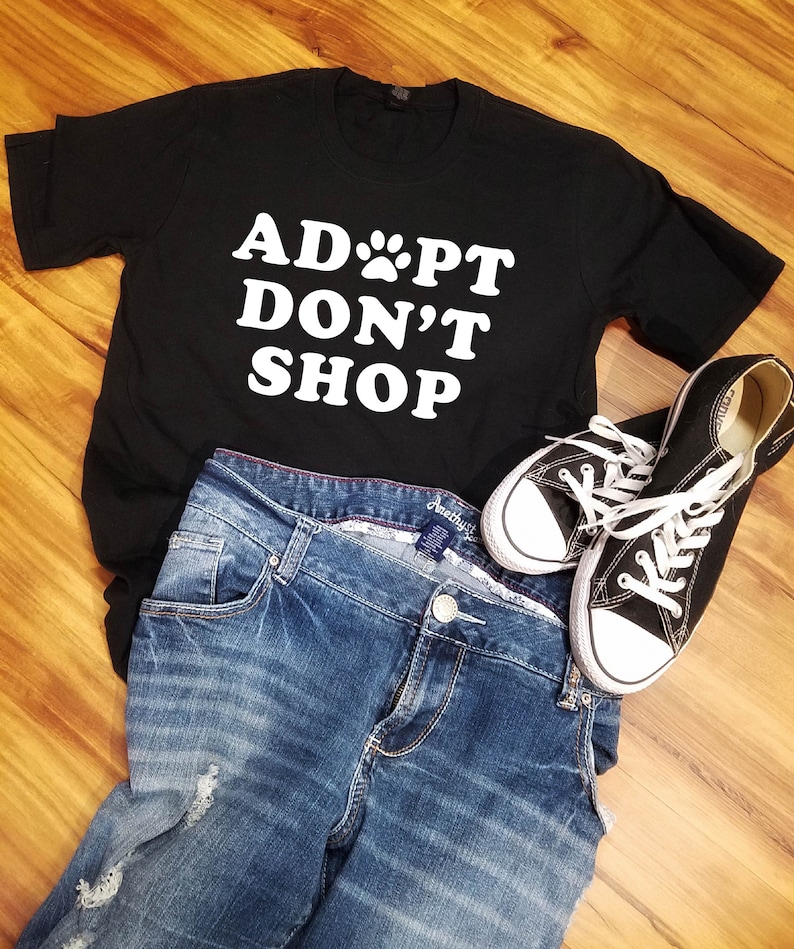 K&ouml;nnte beinhalten: Ein schwarzes T-Shirt mit dem wei&szlig;en Text "ADOPT DON'T SHOP", kombiniert mit blauen Jeans und schwarzen Canvas-Sneakern mit wei&szlig;en Schn&uuml;rsenkeln. Die Artikel liegen auf einer Holzoberfl&auml;che.
