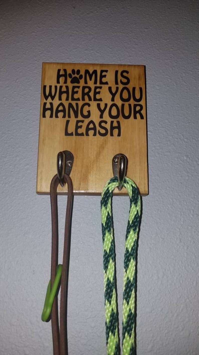 Op de afbeelding: Een houten bord met de tekst "Home is where you hang your leash" met twee haken en daaraan hangende leibanden. Een leiband is van bruin leer en de andere is groen en geel.