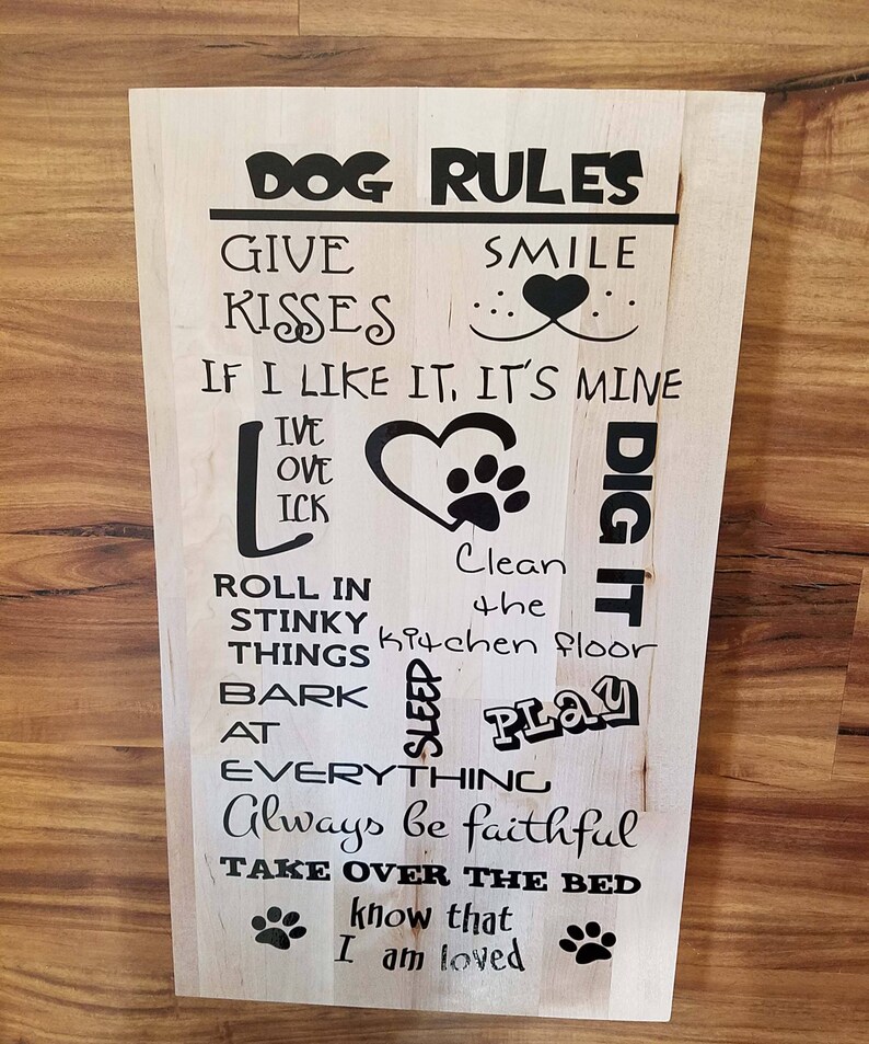 Op de afbeelding: Een houten bord met zwarte tekst die "DOG RULES" luidt en een reeks regels voor honden opsomt, waaronder "Give Kisses", "Smile", "If I like it, it's mine", "Roll in stinky things", "Bark at everything", "Always be faithful" en "Take over the bed". Het bord bevat ook poot afdrukken en een hart.