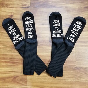 May include: A pair of black socks with white text. One sock says "I just want to drink whiskey" and the other says "And hang out with my cat".