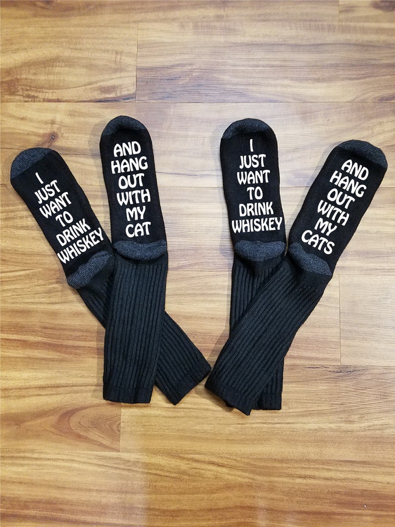May include: A pair of black socks with white text. One sock says "I just want to drink whiskey" and the other says "And hang out with my cat".