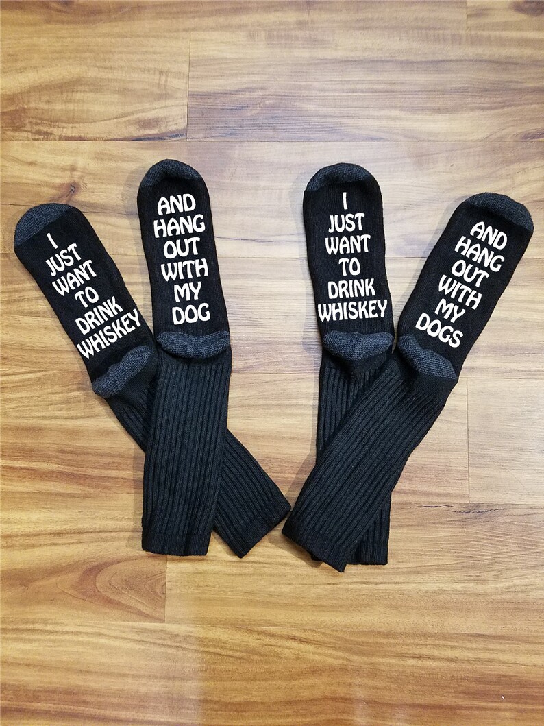 May include: Four black socks with white text. The socks have different sayings on them, including "I just want to drink whiskey", "And hang out with my dog", "I just want to drink whiskey", and "And hang out with my dogs".