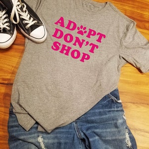 K&ouml;nnte beinhalten: Ein graues T-Shirt mit dem pinkfarbenen Text "ADOPT DON'T SHOP", kombiniert mit blauen Jeans und schwarzen Canvas-Sneakers mit wei&szlig;en Schn&uuml;rsenkeln und Sohlen. Das Outfit liegt auf einer Holzoberfl&auml;che.