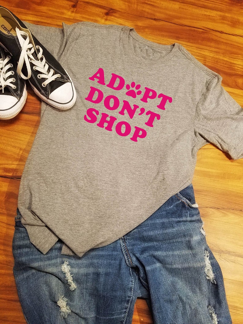 K&ouml;nnte beinhalten: Ein graues T-Shirt mit dem pinkfarbenen Text "ADOPT DON'T SHOP", kombiniert mit blauen Jeans und schwarzen Canvas-Sneakers mit wei&szlig;en Schn&uuml;rsenkeln und Sohlen. Das Outfit liegt auf einer Holzoberfl&auml;che.