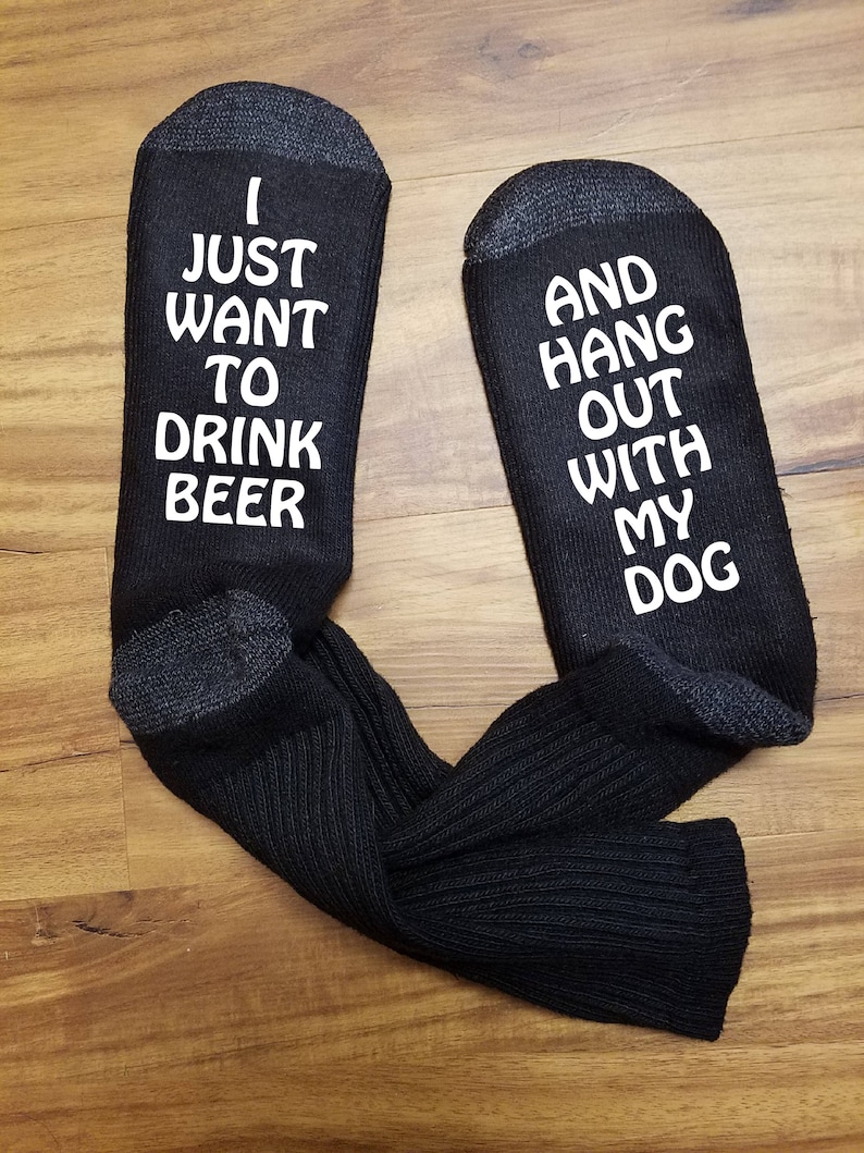 May include: A pair of black socks with white text. One sock reads "I just want to drink beer" and the other sock reads "and hang out with my dog".