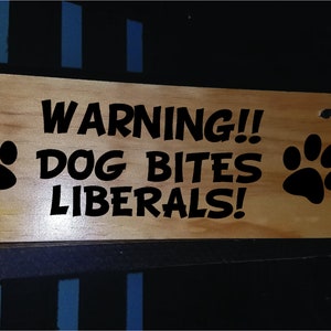 Puede incluir: Un letrero de madera con texto negro que dice "WARNING!! DOG BITES LIBERALS!" con una huella de pata negra a cada lado.