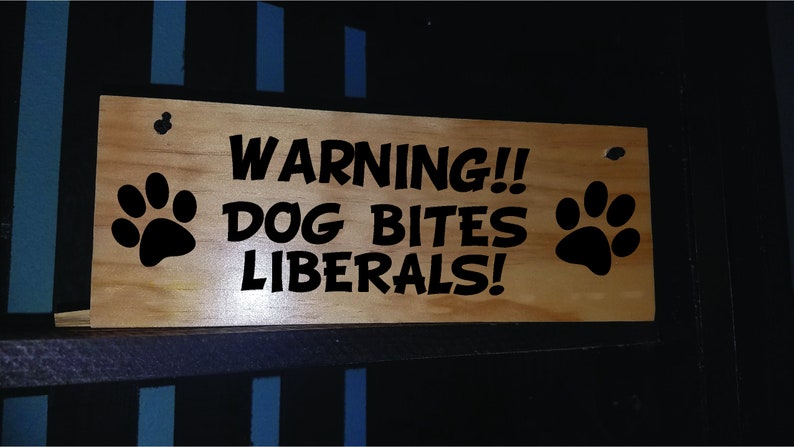 Puede incluir: Un letrero de madera con texto negro que dice "WARNING!! DOG BITES LIBERALS!" con una huella de pata negra a cada lado.