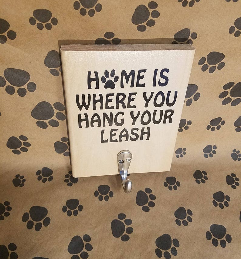 Op de afbeelding: Een houten bord met de tekst "Home is where you hang your leash" en een metalen haak. Het bord is op een bruine achtergrond met poot afdrukken.