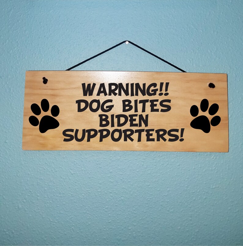 Puede incluir: Un letrero de madera con texto negro que dice "WARNING!! DOG BITES BIDEN SUPPORTERS!" con una huella de pata negra a cada lado.