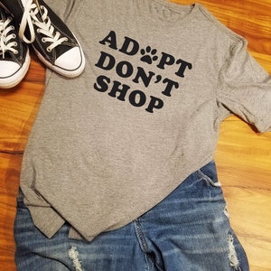 K&ouml;nnte beinhalten: Ein graues T-Shirt mit dem schwarzen Text "ADOPT DON'T SHOP", kombiniert mit blauen Jeans und schwarzen Canvas-Sneakern mit wei&szlig;en Schn&uuml;rsenkeln. Die Jeans haben Distressed-Details.