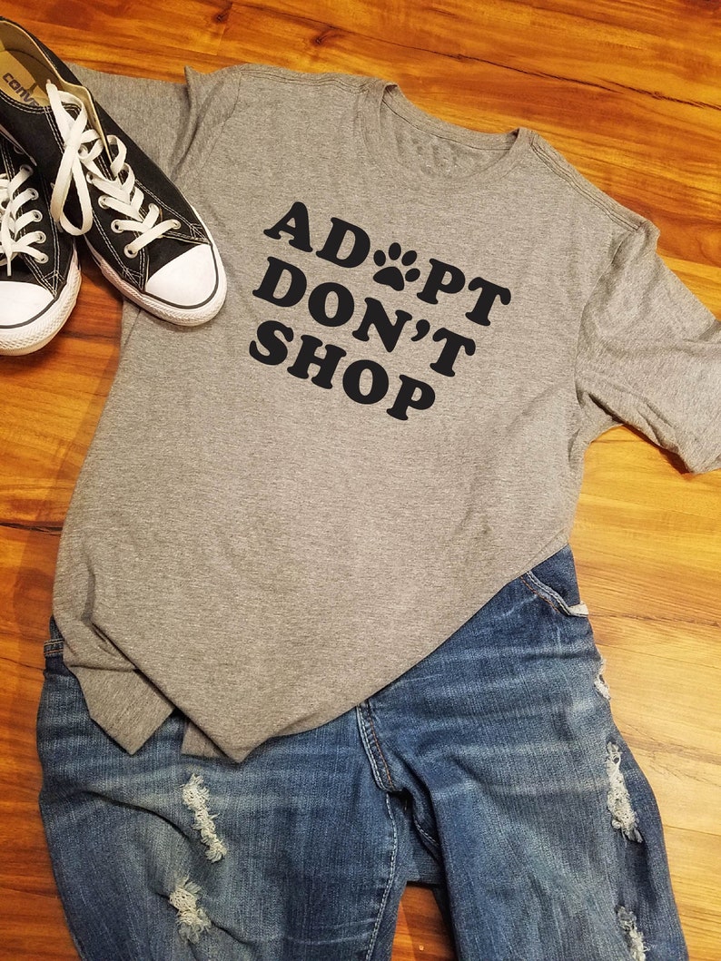 K&ouml;nnte beinhalten: Ein graues T-Shirt mit dem schwarzen Text "ADOPT DON'T SHOP", kombiniert mit blauen Jeans und schwarzen Canvas-Sneakern mit wei&szlig;en Schn&uuml;rsenkeln. Die Jeans haben Distressed-Details.