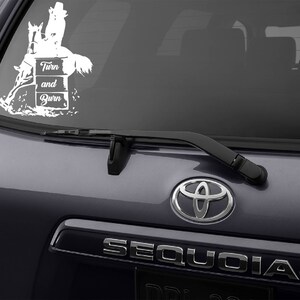 May include: White vinyl decal of a horse and rider jumping over a barrel with the text "Turn and Burn". The decal is on the back window of a gray SUV.