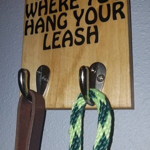 Op de afbeelding: Een houten bord met de tekst "Home is where you hang your leash" met twee haken en daaraan hangende leibanden. Een leiband is van bruin leer en de andere is van groen en geel touw.