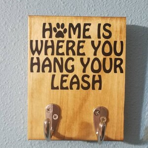 Op de afbeelding: Een houten bord met de tekst "Home is where you hang your leash" en een pootafdruk. Het bord heeft twee zilveren haken.