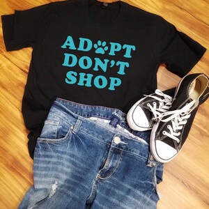 K&ouml;nnte beinhalten: Schwarzes T-Shirt mit dem t&uuml;rkisfarbenen Text "ADOPT DON'T SHOP", kombiniert mit blauen Jeans und schwarzen Canvas-Sneakers mit wei&szlig;en Schn&uuml;rsenkeln. Die Artikel sind auf einer Holzoberfl&auml;che angeordnet.