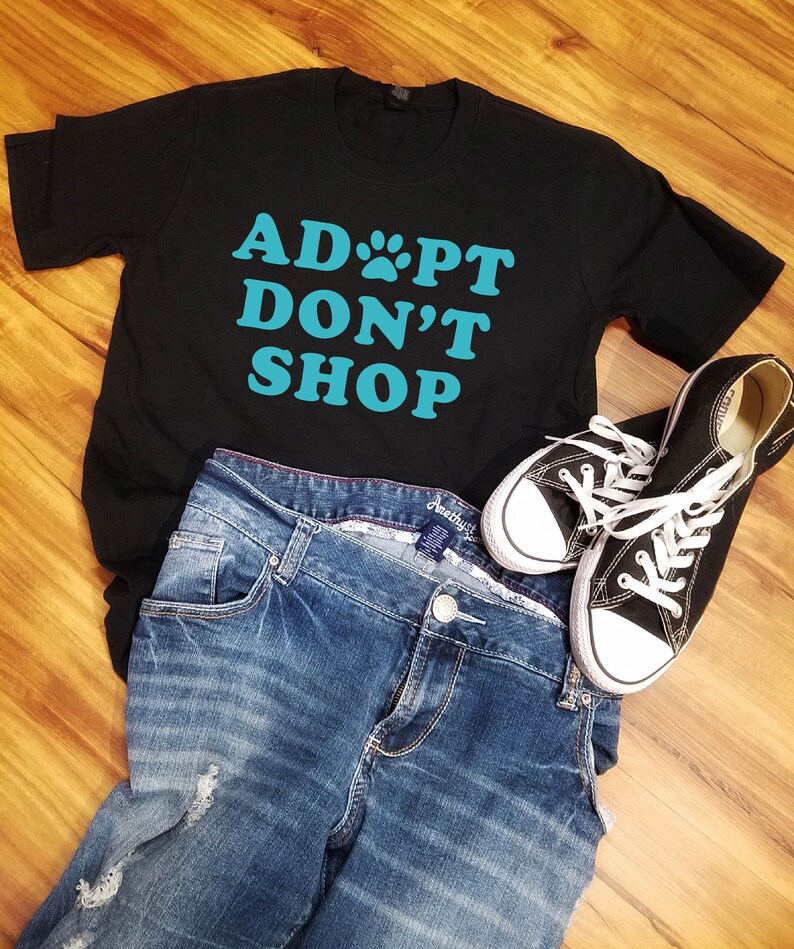 K&ouml;nnte beinhalten: Schwarzes T-Shirt mit dem t&uuml;rkisfarbenen Text "ADOPT DON'T SHOP", kombiniert mit blauen Jeans und schwarzen Canvas-Sneakers mit wei&szlig;en Schn&uuml;rsenkeln. Die Artikel sind auf einer Holzoberfl&auml;che angeordnet.