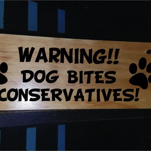 Puede incluir: Un letrero de madera con texto negro que dice "WARNING!! DOG BITES CONSERVATIVES!" con una huella de pata a cada lado.