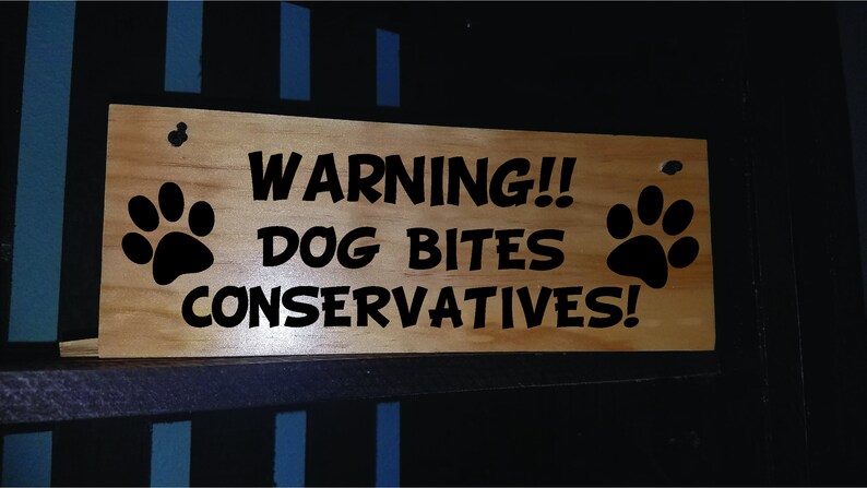 Puede incluir: Un letrero de madera con texto negro que dice "WARNING!! DOG BITES CONSERVATIVES!" con una huella de pata a cada lado.