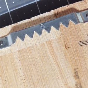 Op de afbeelding: Een close-up van een zilveren zaagtand-beeld ophanghaak bevestigd aan een houten oppervlak.