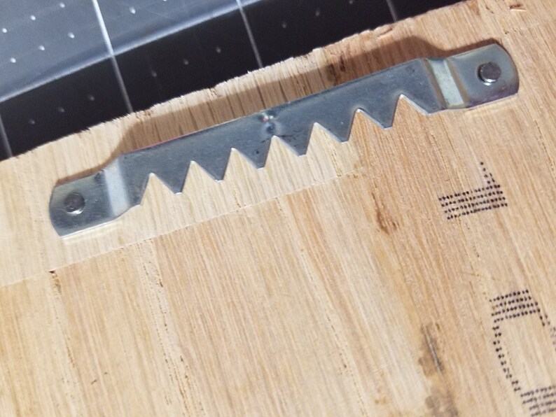 Op de afbeelding: Een close-up van een zilveren zaagtand-beeld ophanghaak bevestigd aan een houten oppervlak.
