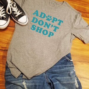 K&ouml;nnte beinhalten: Ein graues T-Shirt mit dem t&uuml;rkisfarbenen Text "ADOPT DON'T SHOP", wobei ein Pfotenabdruck das "O" ersetzt. Das Bild zeigt blaue Jeans und schwarze Leinenschuhe mit wei&szlig;en Schn&uuml;rsenkeln.