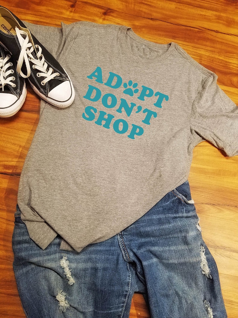 K&ouml;nnte beinhalten: Ein graues T-Shirt mit dem t&uuml;rkisfarbenen Text "ADOPT DON'T SHOP", wobei ein Pfotenabdruck das "O" ersetzt. Das Bild zeigt blaue Jeans und schwarze Leinenschuhe mit wei&szlig;en Schn&uuml;rsenkeln.