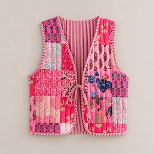 Puede incluir: Un chaleco acolchado rosa con un diseño de patchwork de patrones florales y paisley. El chaleco tiene un forro a rayas y un cierre de lazo. Los bordes están ribeteados con costuras blancas.