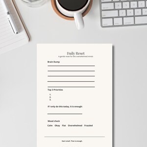 Puede incluir: Un bloc de notas blanco "Daily Reset" con indicaciones para un "brain dump", las principales prioridades y una comprobación del estado de ánimo. Sobre un escritorio blanco, con una taza de café y un teclado.