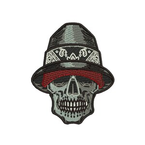 Pode incluir: Um emblema bordado apresentando um crânio cinza claro usando um chapéu fedora cinza escuro. O chapéu é acentuado com uma faixa de padrão paisley em preto e branco e um fino acabamento vermelho ao longo da aba. O design é contornado em fio preto.