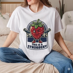 May include: White t-shirt with a cartoon strawberry design, winking and holding guns, with the text "THIS IS A STROBBERY". The person wearing the shirt is also wearing blue jeans.
