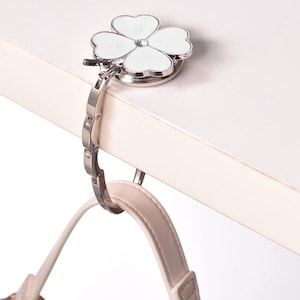 May include: A silver purse hook with a four-leaf clover design. The clover has white enamel petals and a small crystal center. The hook is attached to a silver chain and is holding a light pink purse strap.