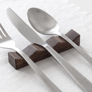 Könnte beinhalten: Ein Besteckset aus Edelstahl, bestehend aus einer Gabel, einem Messer und einem Löffel, das auf einer dunkelbraunen hölzernen Besteckablage ruht. Das Besteck zeichnet sich durch eine glatte, matte Oberfläche und schlichte, klare Linien aus.