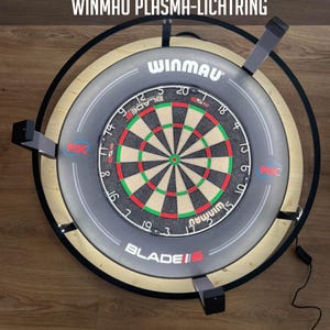 May include: A Winmau dartboard with a gray surround and a black ring light. The dartboard features a classic design with red, green, and black segments. Text on the surround reads "WINMAU" and "BLADE 6". The image also includes text that reads "AUTO SCORING KAMERA SYSTEM FÜR WINMAU PLASMA-LICHTRING".