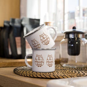 Pode incluir: Duas canecas de esmalte branco empilhadas sobre uma esteira tecida. Cada caneca apresenta uma ilustração de coruja marrom. As canecas têm bordas e alças prateadas. Uma chaleira de vidro e sacos de grãos de café estão no fundo.