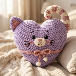 May include: A heart-shaped crochet pillow in lavender, featuring a cat face with black eyes, a cream-colored muzzle, pink cheeks, and whiskers. It has a striped pink and cream tail, a pink bow tied around the middle, and two small ears.