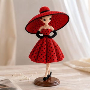 May include: A crocheted doll wearing a red dress with black polka dots, a wide-brimmed red hat with a black trim, and black gloves. The doll has a light beige complexion, a pearl-like necklace, and black high-heeled shoes, standing on a round wooden base.
