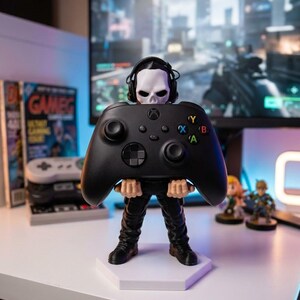 May include: A black Xbox controller held by a figurine with a skull head wearing headphones. The figurine is standing on a white hexagonal base. The background includes a computer monitor displaying a video game and other gaming accessories.