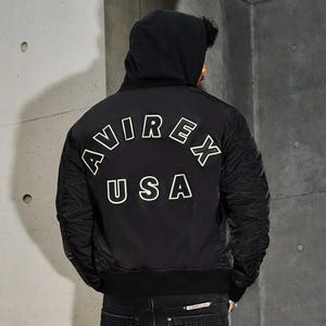 K&ouml;nnte beinhalten: Schwarze Bomberjacke mit Kapuze. Auf der R&uuml;ckseite der Jacke befindet sich der wei&szlig;e Schriftzug "AVIREX USA" in einem geschwungenen Design. Die Jacke hat einen gerippten Saum und B&uuml;ndchen. Das Model tr&auml;gt dunkle Jeans.