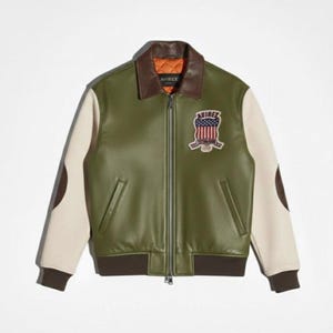Op de afbeelding: Een olijfgroene jas met cr&egrave;mekleurige mouwen, een bruine leren kraag en bruine geribbelde boorden. De jas is voorzien van een ritssluiting aan de voorzijde, twee zijzakken en een geborduurde patch op de borst met de Amerikaanse vlag en tekst.