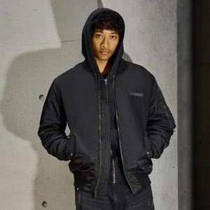 K&ouml;nnte beinhalten: Schwarze Bomberjacke mit Kapuze, Rei&szlig;verschluss und Seitentaschen. Die Jacke hat einen kleinen rechteckigen Aufn&auml;her auf der Brust. Die Person tr&auml;gt einen schwarzen Kapuzenpullover und eine schwarze Jeans.