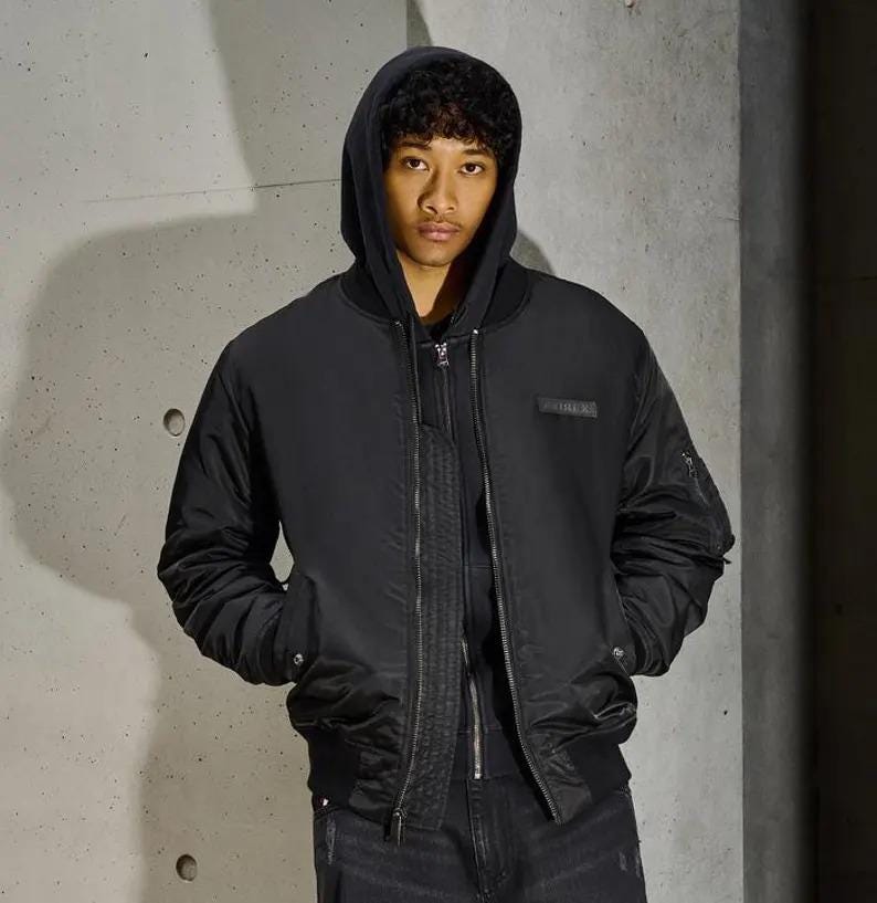 K&ouml;nnte beinhalten: Schwarze Bomberjacke mit Kapuze, Rei&szlig;verschluss und Seitentaschen. Die Jacke hat einen kleinen rechteckigen Aufn&auml;her auf der Brust. Die Person tr&auml;gt einen schwarzen Kapuzenpullover und eine schwarze Jeans.
