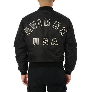 K&ouml;nnte beinhalten: Schwarze Bomberjacke mit wei&szlig;em Schriftzug "AVIREX USA" auf dem R&uuml;cken. Die Jacke hat einen gerippten Kragen, B&uuml;ndchen und Saum. Sie verf&uuml;gt &uuml;ber eine Rei&szlig;verschlusstasche am linken &Auml;rmel.