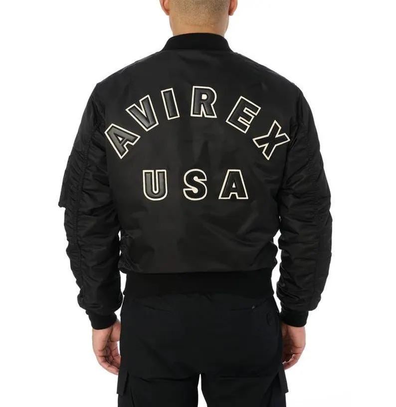 K&ouml;nnte beinhalten: Schwarze Bomberjacke mit wei&szlig;em Schriftzug "AVIREX USA" auf dem R&uuml;cken. Die Jacke hat einen gerippten Kragen, B&uuml;ndchen und Saum. Sie verf&uuml;gt &uuml;ber eine Rei&szlig;verschlusstasche am linken &Auml;rmel.
