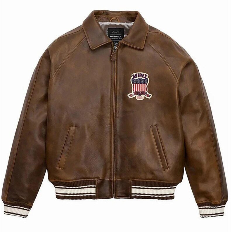 May include: A brown leather jacket featuring a front zipper closure, a pointed collar, and two side pockets. The chest displays a patch with the text AVIREX and a shield graphic containing the American flag. The cuffs and hem have white striped trim.