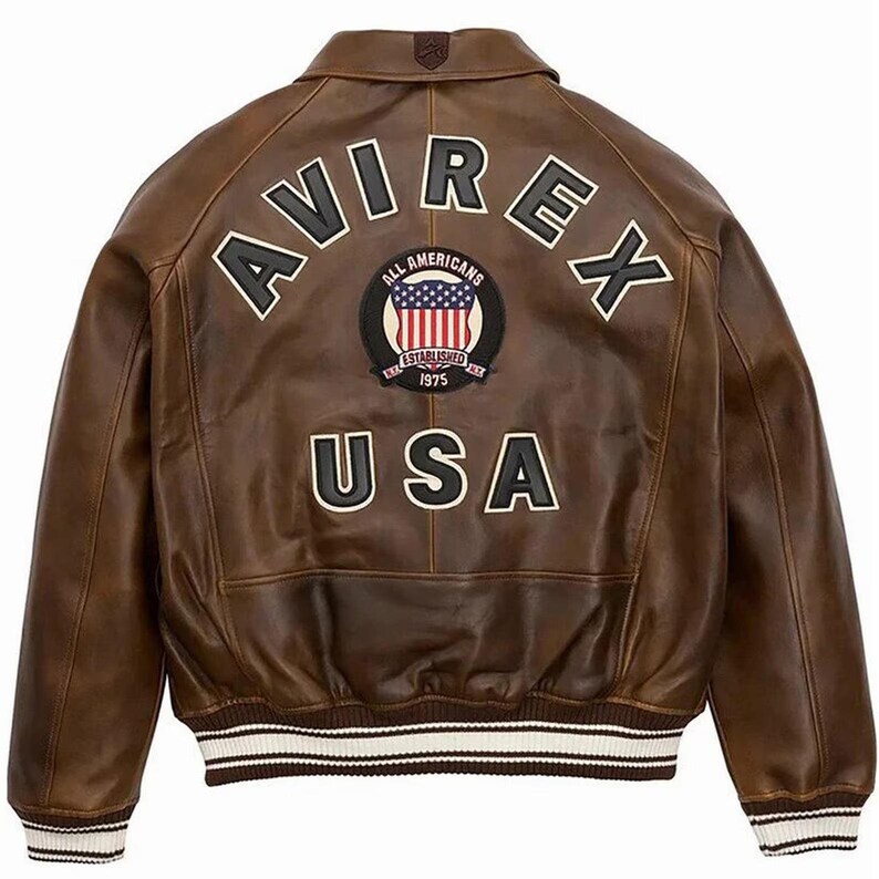 May include: A brown leather bomber jacket featuring the text AVIREX in black and white lettering across the upper back. Below this, a circular patch displays the text ALL AMERICANS ESTABLISHED 1975 with a US flag. The word USA is printed below.