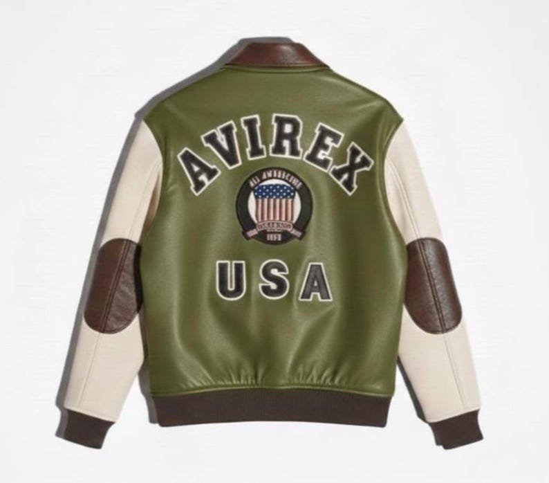 Op de afbeelding: Een olijfgroen varsity-jack met cr&egrave;mekleurige mouwen, bruine leren elleboogstukken en een bruine kraag en zoom. Op de achterkant staat de tekst AVIREX en USA in zwarte letters, samen met een ronde badge met een afbeelding van een Amerikaanse vlag.