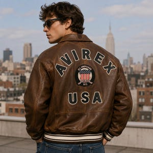 May include: A brown leather bomber jacket featuring the text AVIREX in a curved arch above a circular patch with an American flag design and the text ALL AMERICANS ESTABLISHED 1975. The word USA is printed below the patch in large black letters.